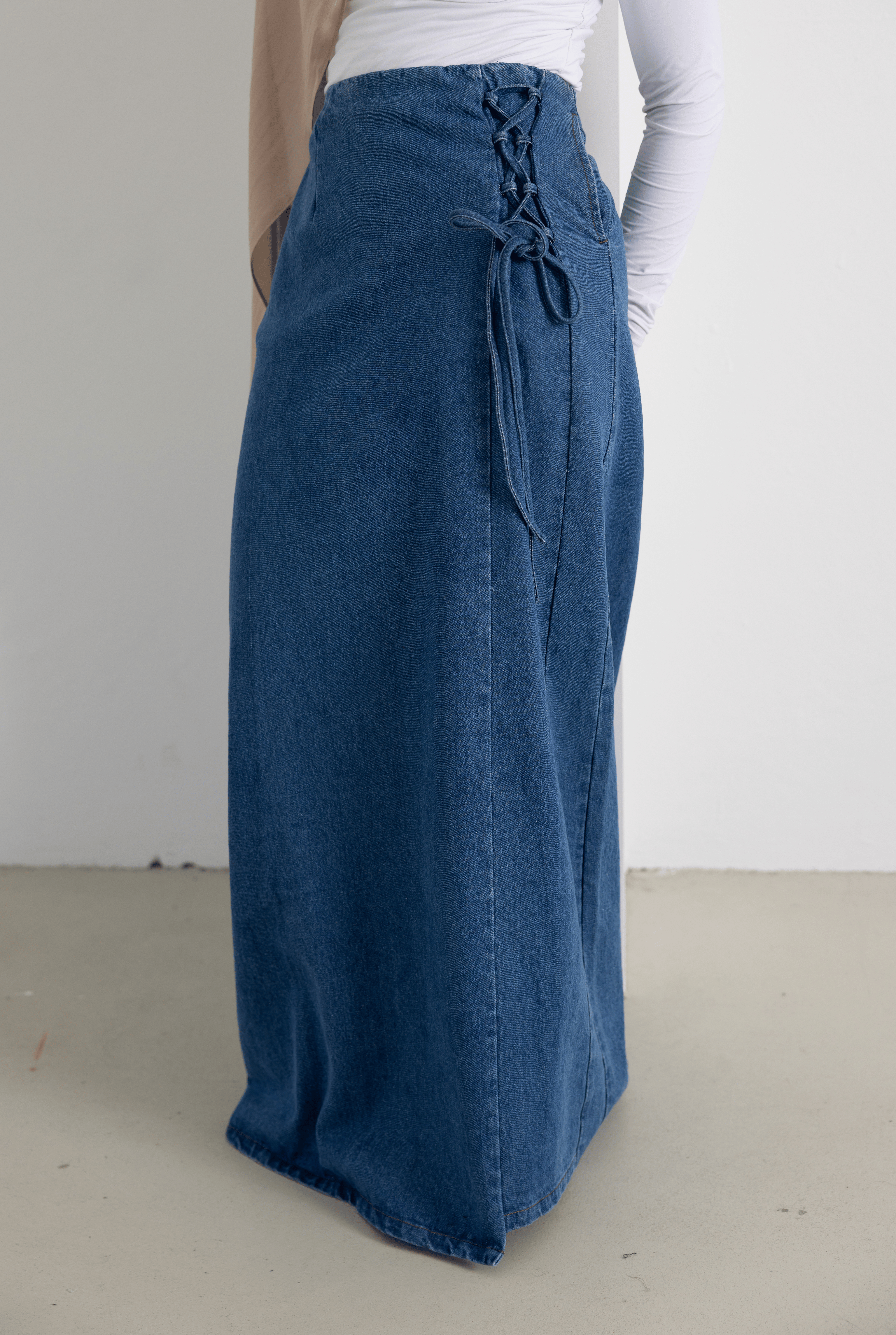 Close-up of the Lace Denim Skirt in mid-wash denim blue, featuring elegant lace-up side detailing and a structured longline silhouette. Made from 100% cotton, this modest denim skirt offers a comfortable fit with a clean front seam and timeless design, perfect for everyday styling and elevated casual looks.