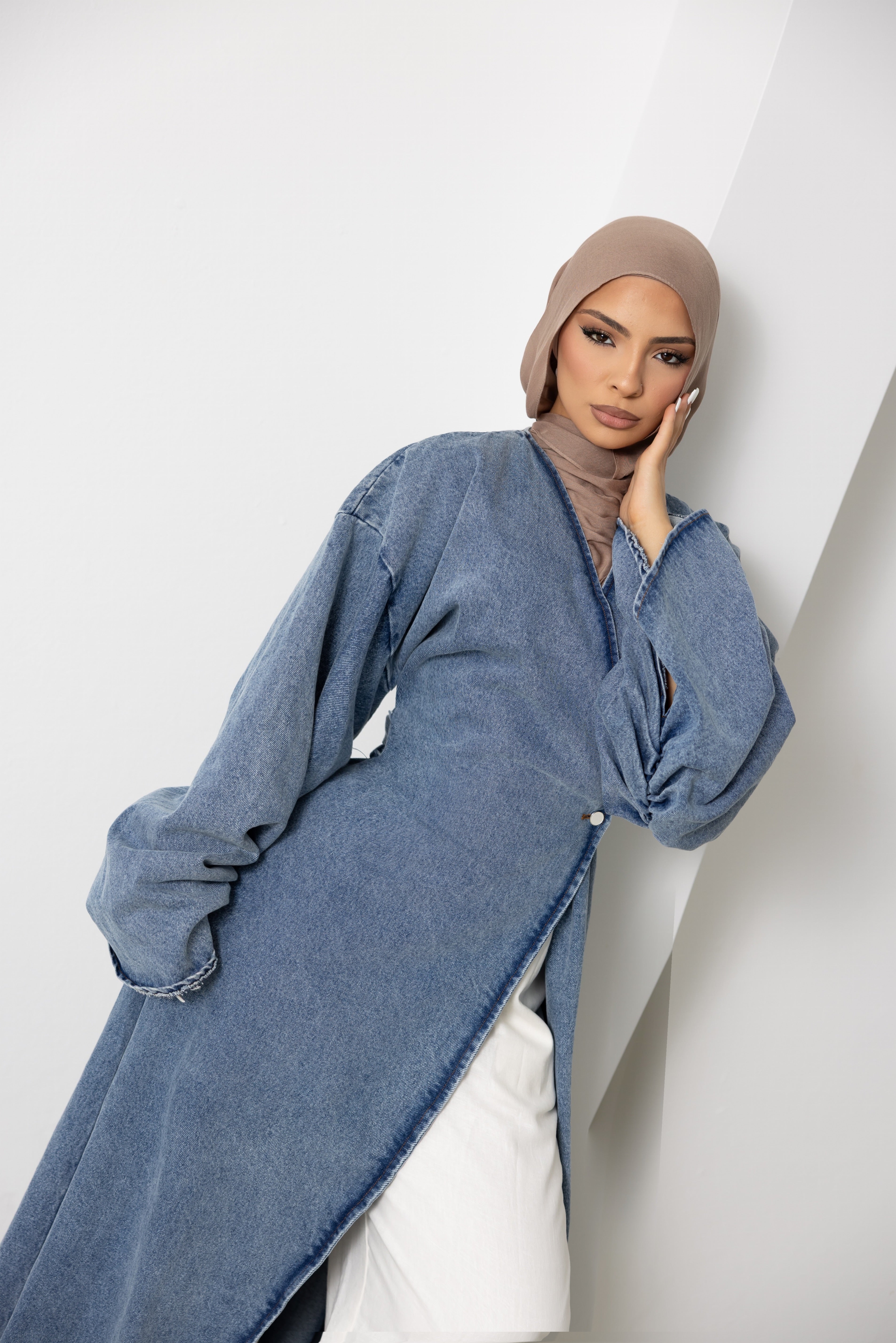 Close-up portrait of a model wearing the Orlena Signature Denim full-length wrap jacket in soft blue 100% cotton denim fabric. Features a flattering wrap-style front, flowing full-length silhouette, wide statement sleeves, and adjustable back tie waist for an elegant fit. Timeless modest fashion layering piece designed for effortless everyday styling and versatile wardrobe wear.