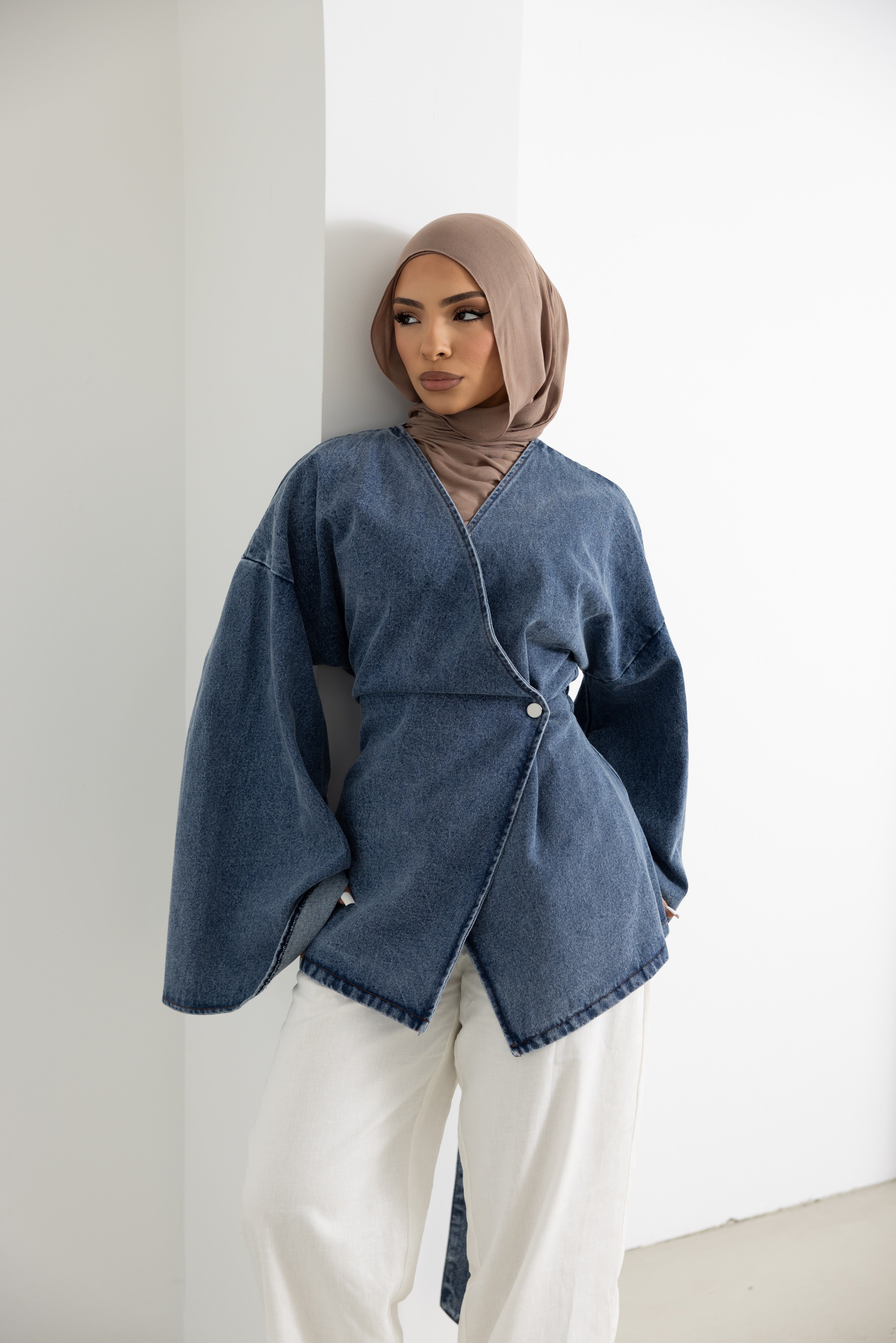 Model wearing the Orlena Signature Denim Mini jacket in premium blue denim-look cotton fabric. Features a wrap-style front, statement wide sleeves, mini-length silhouette, and adjustable back tie for a flattering fit. Modern modest fashion jacket styled with white wide-leg pants, perfect for layering and everyday elevated casual wear.