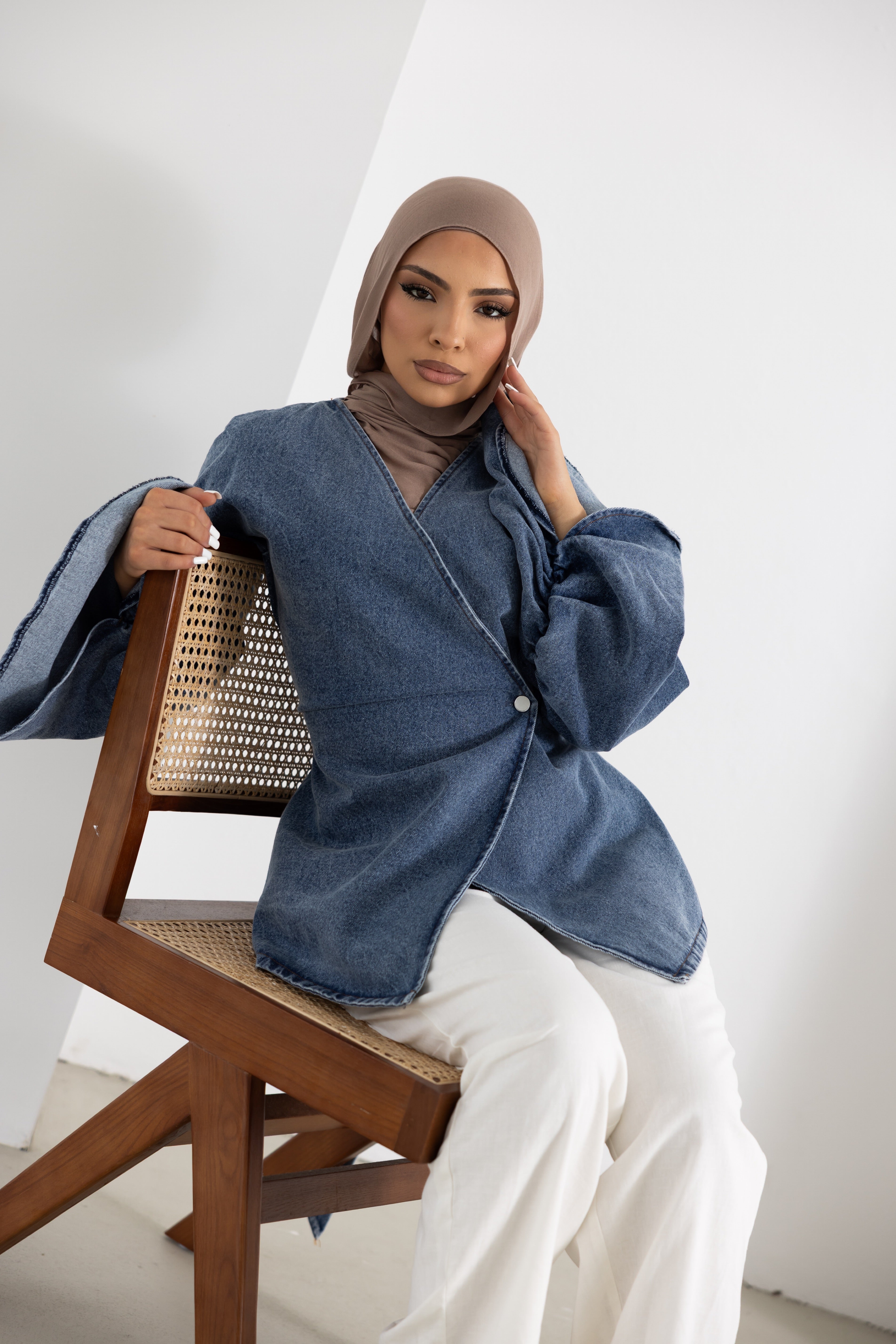Close-up lifestyle image of a model wearing the Orlena Signature Denim Mini jacket in blue denim-look 100% cotton fabric. Features a wrap-style front, wide sleeves, mini-length cut, and adjustable back tie for a flattering silhouette. Styled with white wide-leg pants for an elegant modest fashion look, perfect for layering and everyday wear.