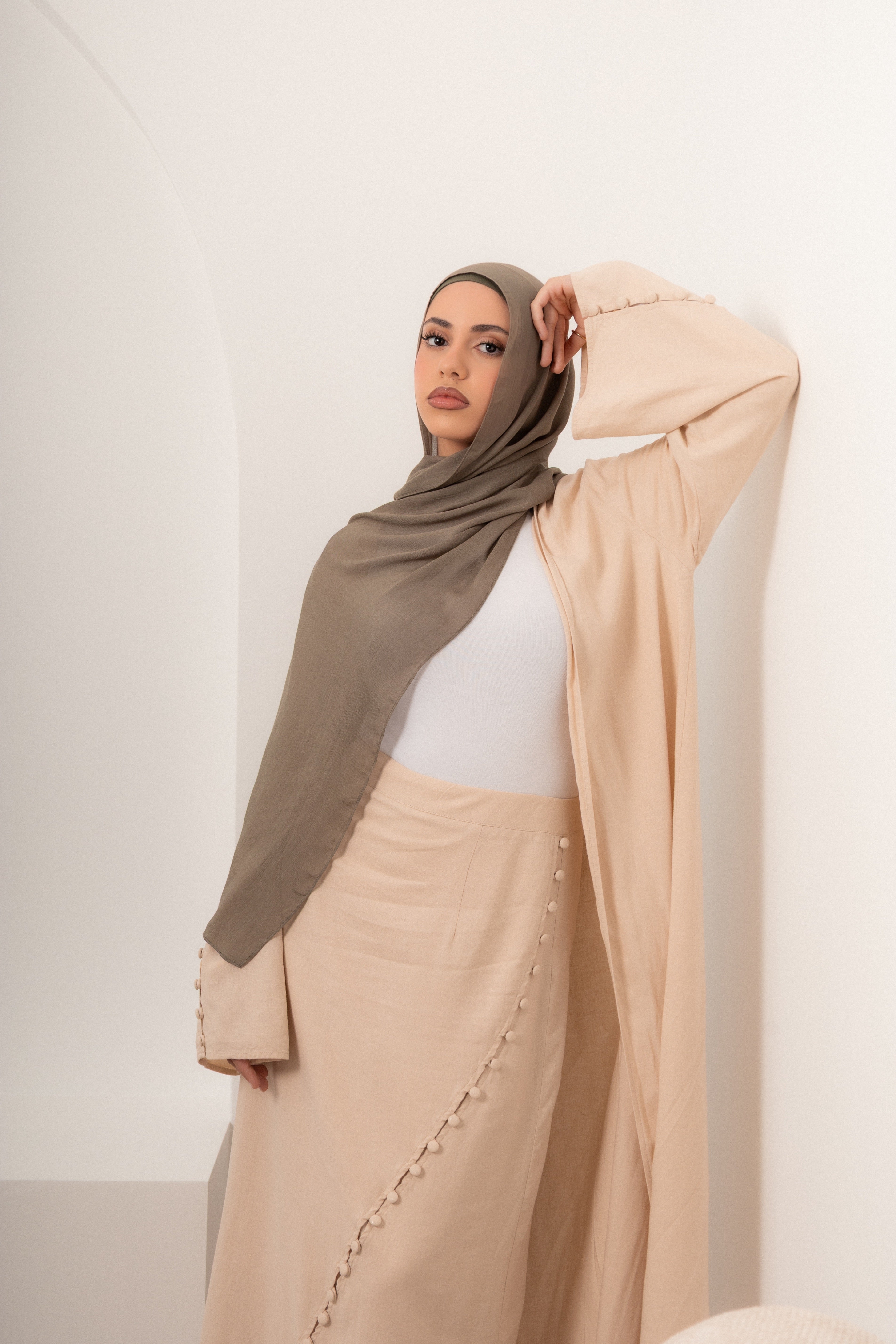 Woman wearing a soft neutral modest outfit with a hijab, featuring a flowing maxi skirt and layered top, styled in a minimalist setting