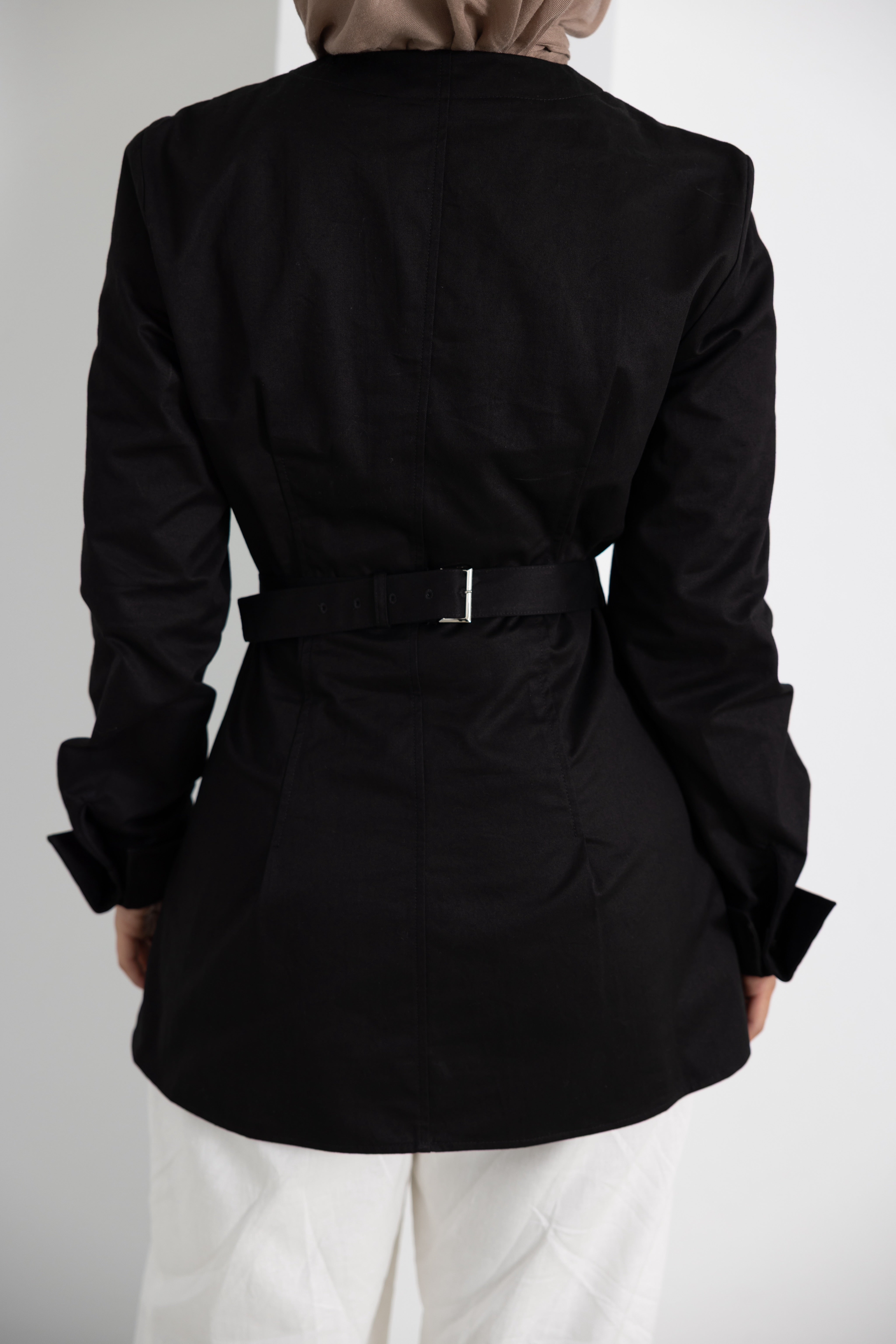 Rear view detail of the Vellisse Top in black, featuring an adjustable back belt that allows the waist to be gently cinched for a flattering and tailored silhouette. Crafted from 100% cotton with a structured yet comfortable fit, showcasing clean panel detailing, long sleeves, and a polished modest design. Styled with white wide-leg pants and a neutral-toned hijab, highlighting the adjustable fit, elegant construction, and versatile everyday wear appeal.