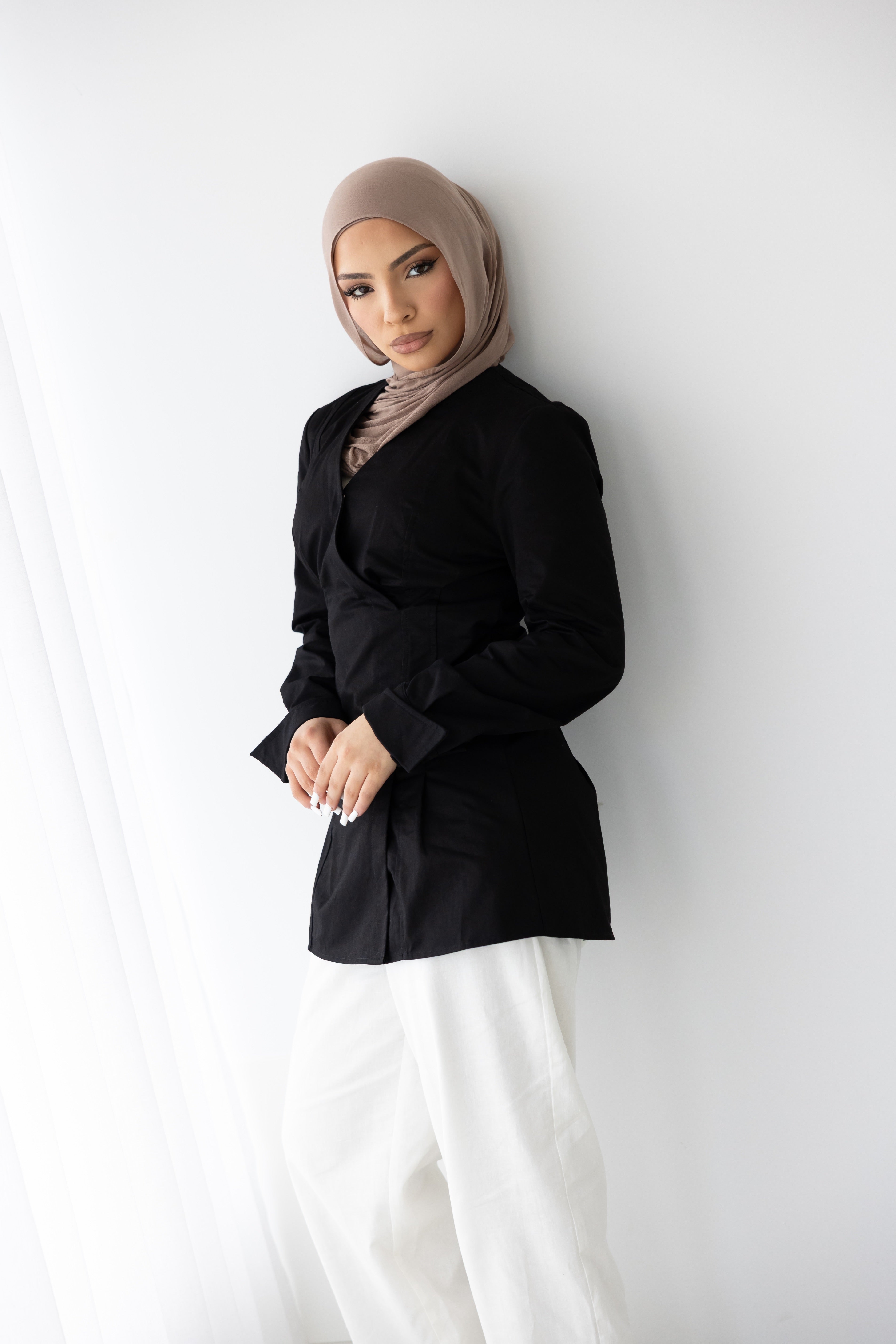 Women's Vellisse Top in black featuring a flattering wrap-style front, long sleeves, and an adjustable back belt for a defined waist silhouette. Made from 100% cotton with a structured yet comfortable fit. Styled with white wide-leg pants, showcasing its versatile modest fashion design that can be dressed up or down for work, casual wear, special occasions, and everyday styling.