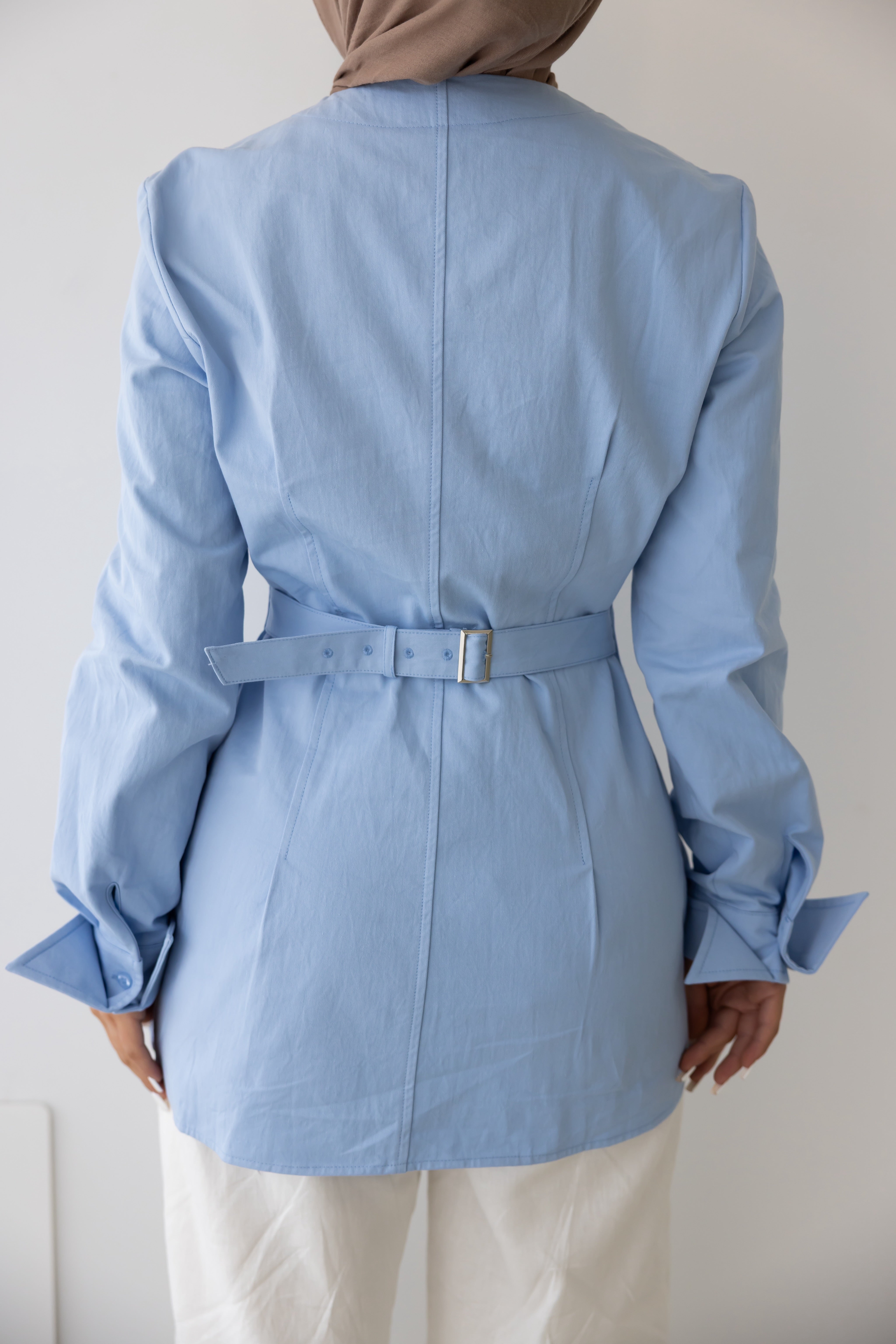 Rear view detail of the Vellisse Top in blue, showcasing the adjustable back belt designed to gently cinch the waist for a flattering and defined silhouette. Crafted from 100% cotton with a structured yet comfortable fit, featuring long sleeves, tailored panel detailing, and a modest design. Styled with white wide-leg pants and a neutral-toned hijab, highlighting the elegant back construction, adjustable fit, and versatile everyday styling.