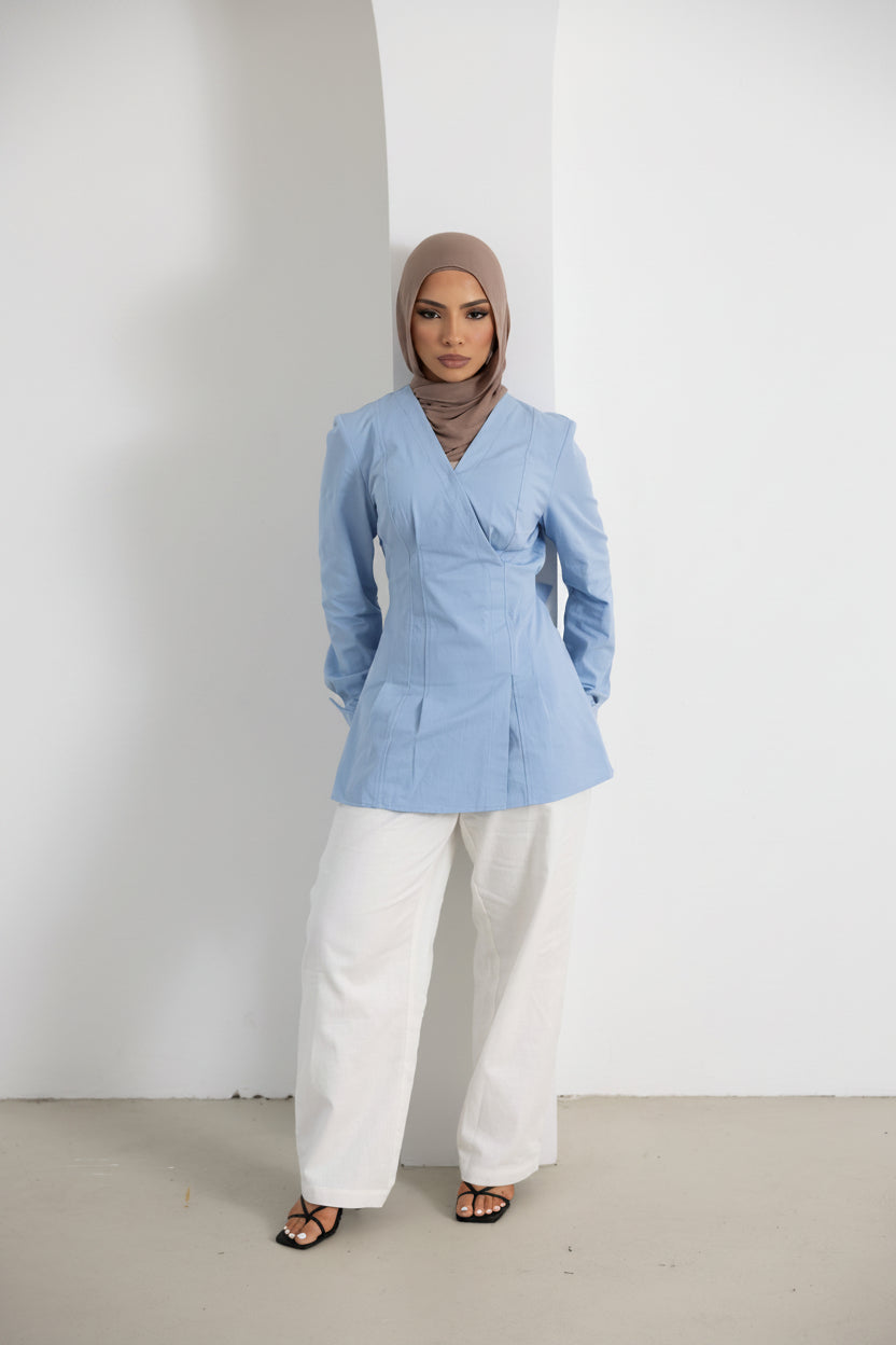 Vellisse Top in blue made from 100% cotton, featuring a flattering wrap-style front, structured silhouette, long sleeves, and an adjustable back belt for a defined waist. Styled with white wide-leg pants and black sandals for a modern modest fashion look. Comfortable and versatile women's cotton top suitable for everyday wear, workwear, and elevated casual styling.