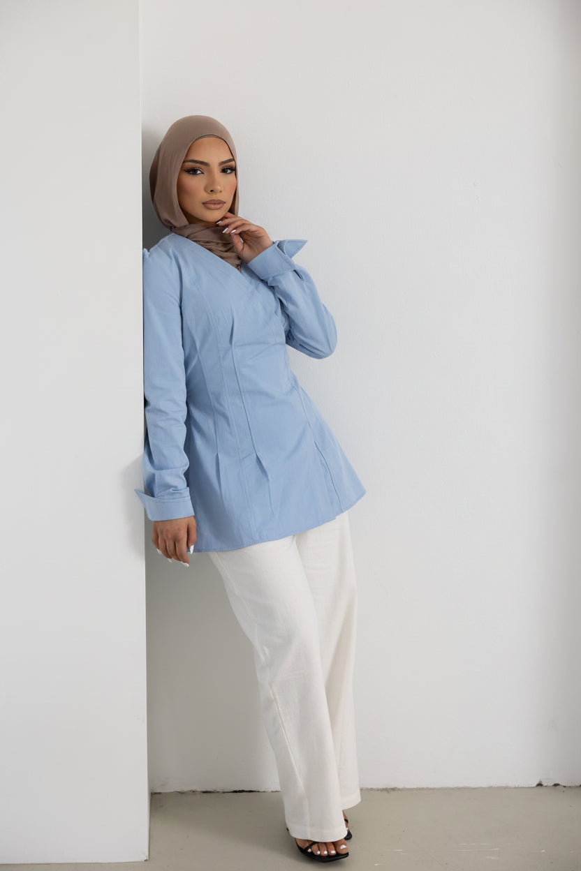 Vellisse Top in blue crafted from 100% cotton, featuring a flattering wrap-style front, adjustable back belt, long sleeves, and a structured tailored silhouette. Styled with white wide-leg pants and black sandals while leaning against a wall, showcasing the elegant modest design and waist-defining fit. A versatile women's cotton top perfect for workwear, smart casual outfits, and everyday modest fashion styling.