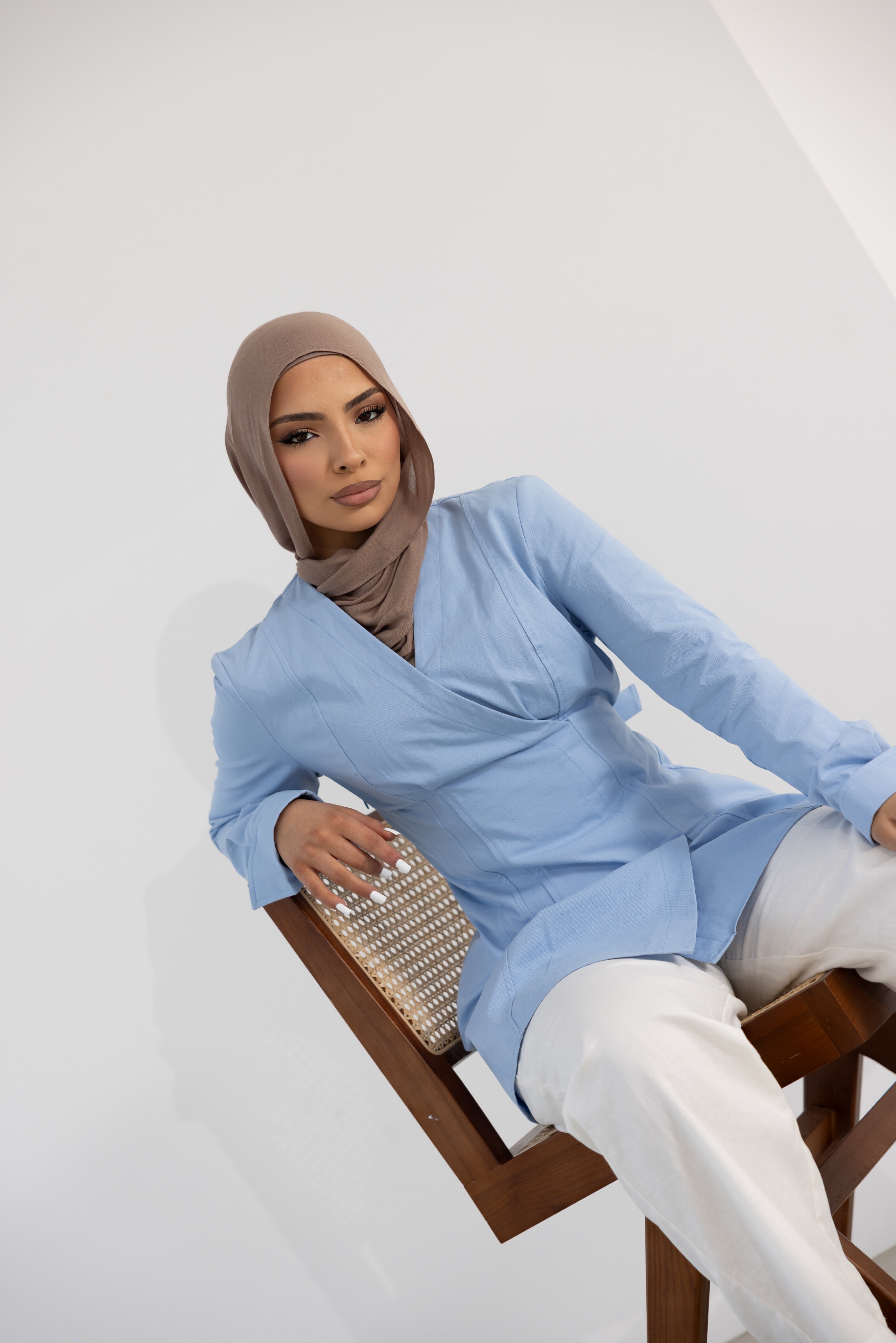 Vellisse Top in blue made from 100% cotton, featuring a flattering wrap-style front, adjustable back belt, and structured tailored fit. Styled with white wide-leg pants while seated on a wooden chair, showcasing the elegant silhouette and modest design. A versatile women's cotton top perfect for everyday wear, office styling, smart casual outfits, and modern modest fashion.