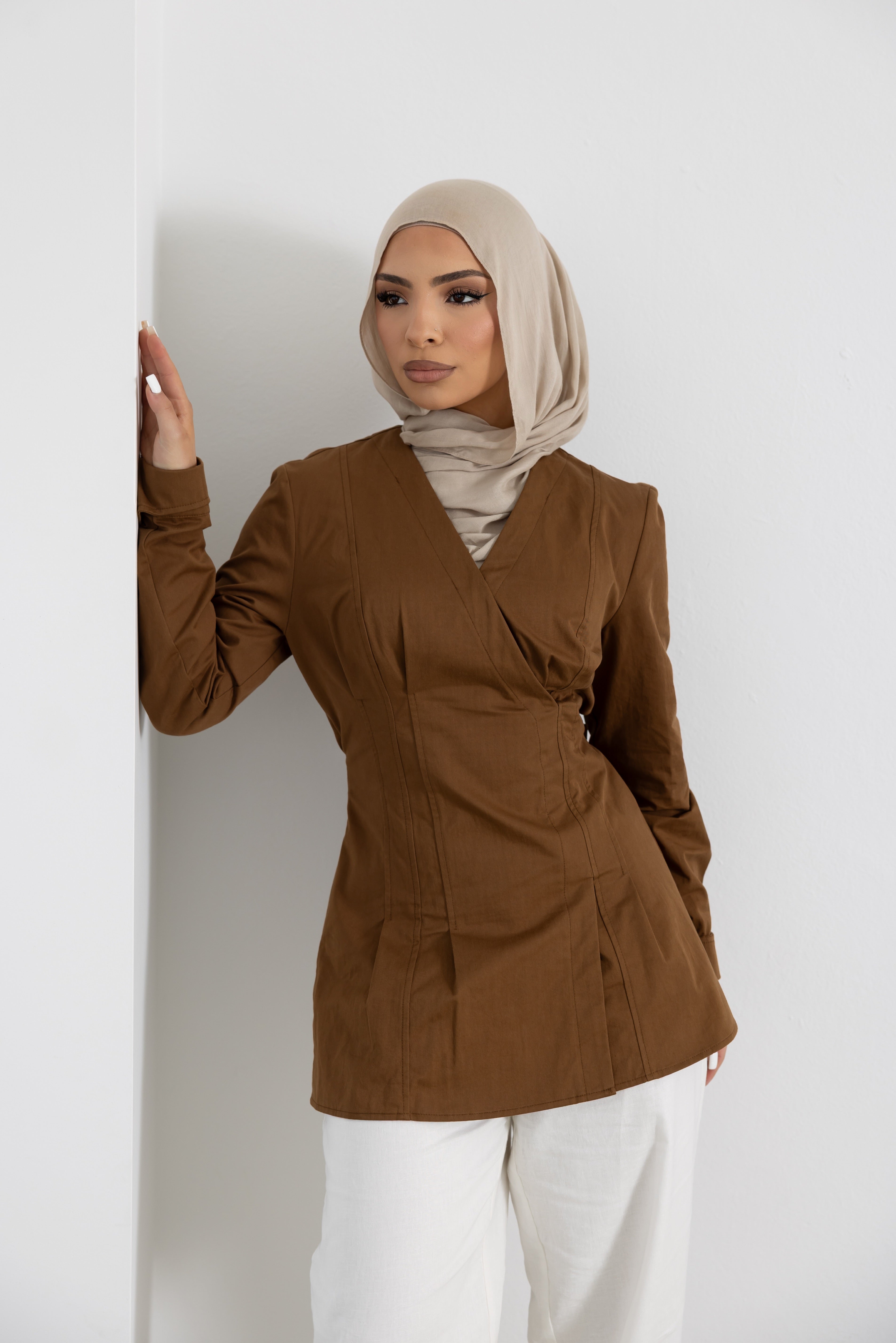Women's Vellisse Top in brown featuring a structured wrap-style front, adjustable back belt for a cinched waist silhouette, and long sleeves. Crafted from 100% cotton for breathable comfort and everyday wear. Styled with white wide-leg pants, showcasing the flattering tailored fit and versatile design suitable for modest fashion, workwear, casual outings, and elevated everyday styling.