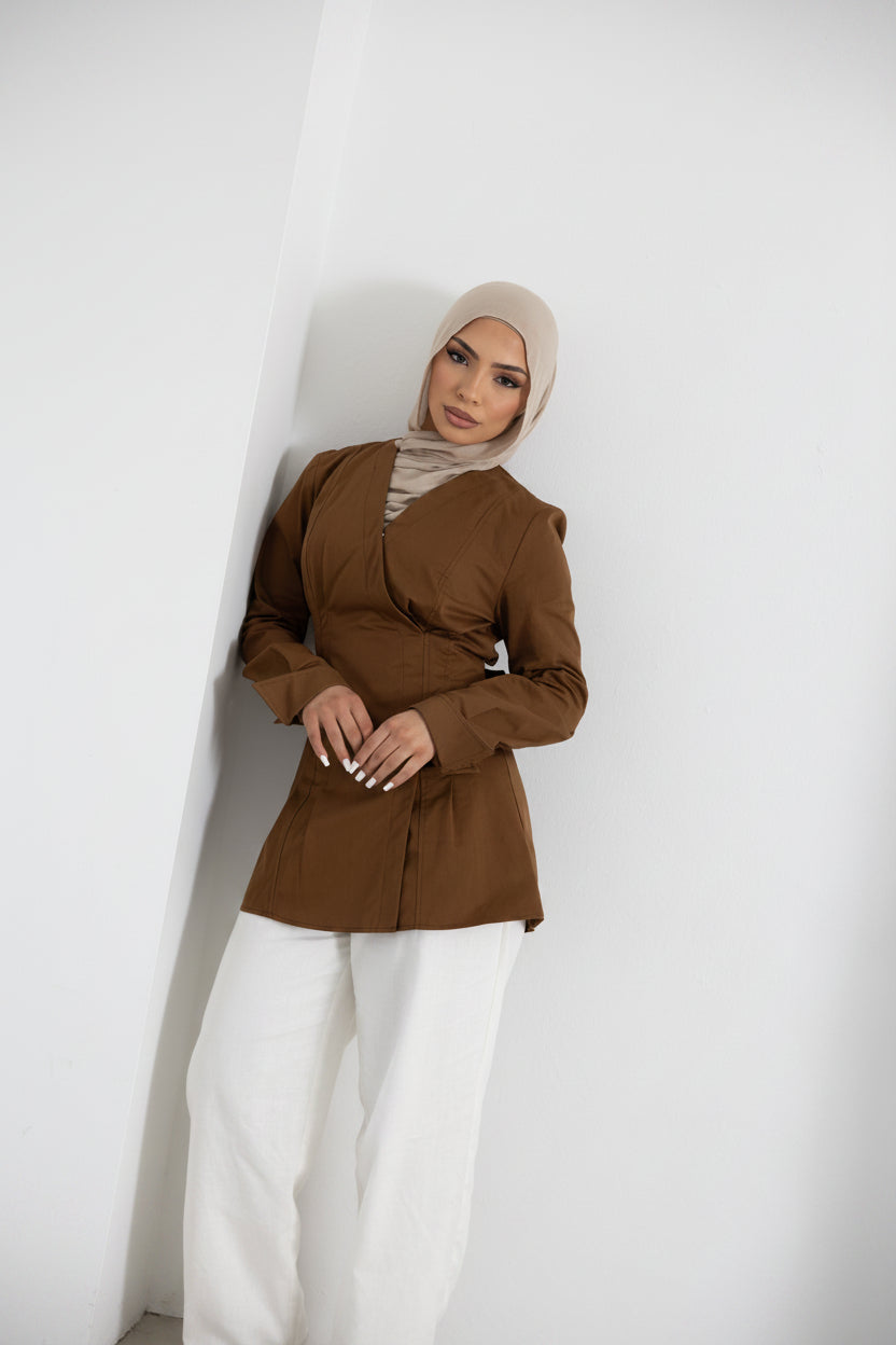 Lifestyle image of a model wearing the Vellisse Top in brown, crafted from 100% cotton with a structured yet comfortable fit. Features a flattering wrap-style front, long sleeves, and an adjustable back belt that creates a defined waist silhouette. Styled with white wide-leg pants for a polished modest fashion look, ideal for both casual styling and elevated everyday wear.