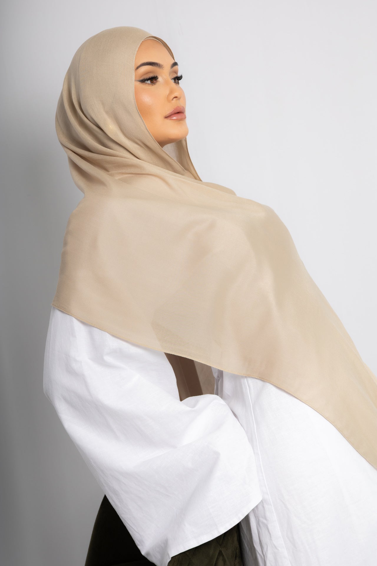 Side profile of a woman wearing a beige hijab styled in an elegant modest fashion look against a clean studio background