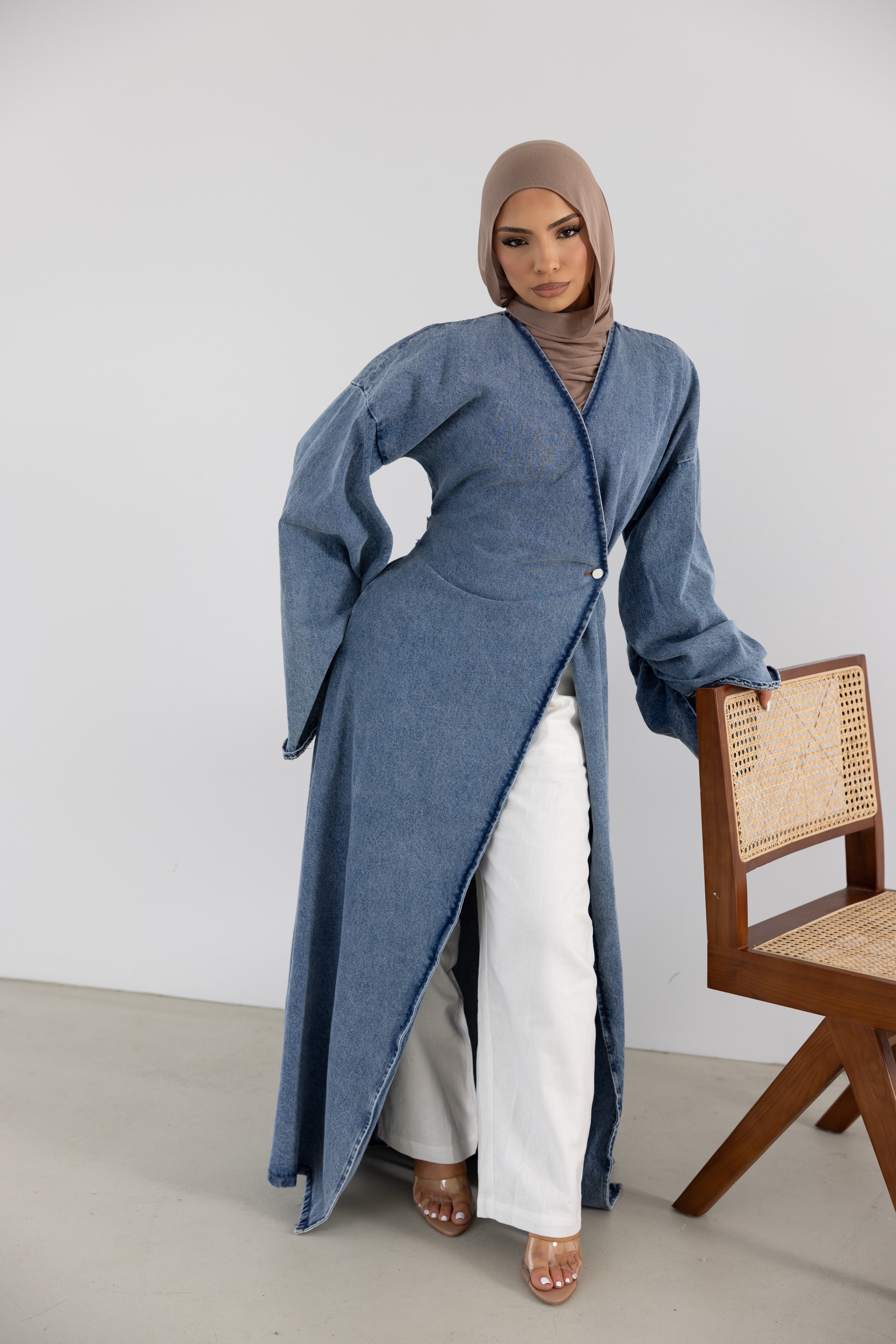 Model wearing the Orlena Signature Denim full-length wrap jacket in blue 100% cotton denim fabric. Features a flattering wrap-style front, full-length silhouette, wide statement sleeves, and adjustable back tie waist for a tailored fit. Elegant modest fashion layering piece styled with white wide-leg pants, designed for effortless everyday wear and timeless wardrobe versatility.
