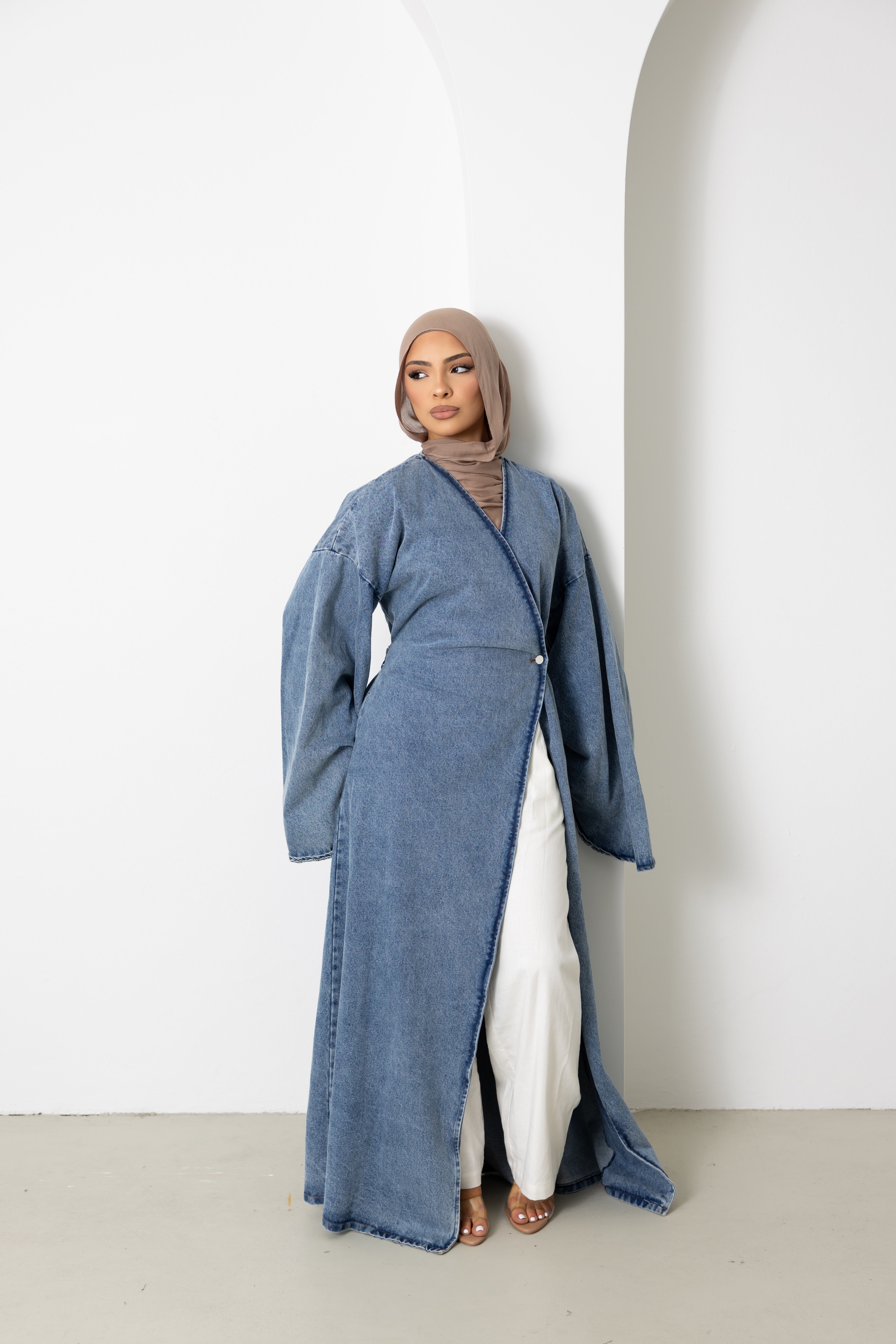 Full-body view of a model wearing the Orlena Signature Denim full-length wrap jacket crafted from 100% cotton denim fabric. Features a wrap-style front, flowing full-length silhouette, wide sleeves, and adjustable back tie waist for a flattering fit. Styled with white wide-leg pants and heels, creating a sophisticated modest fashion look ideal for layering and elevated everyday wear.