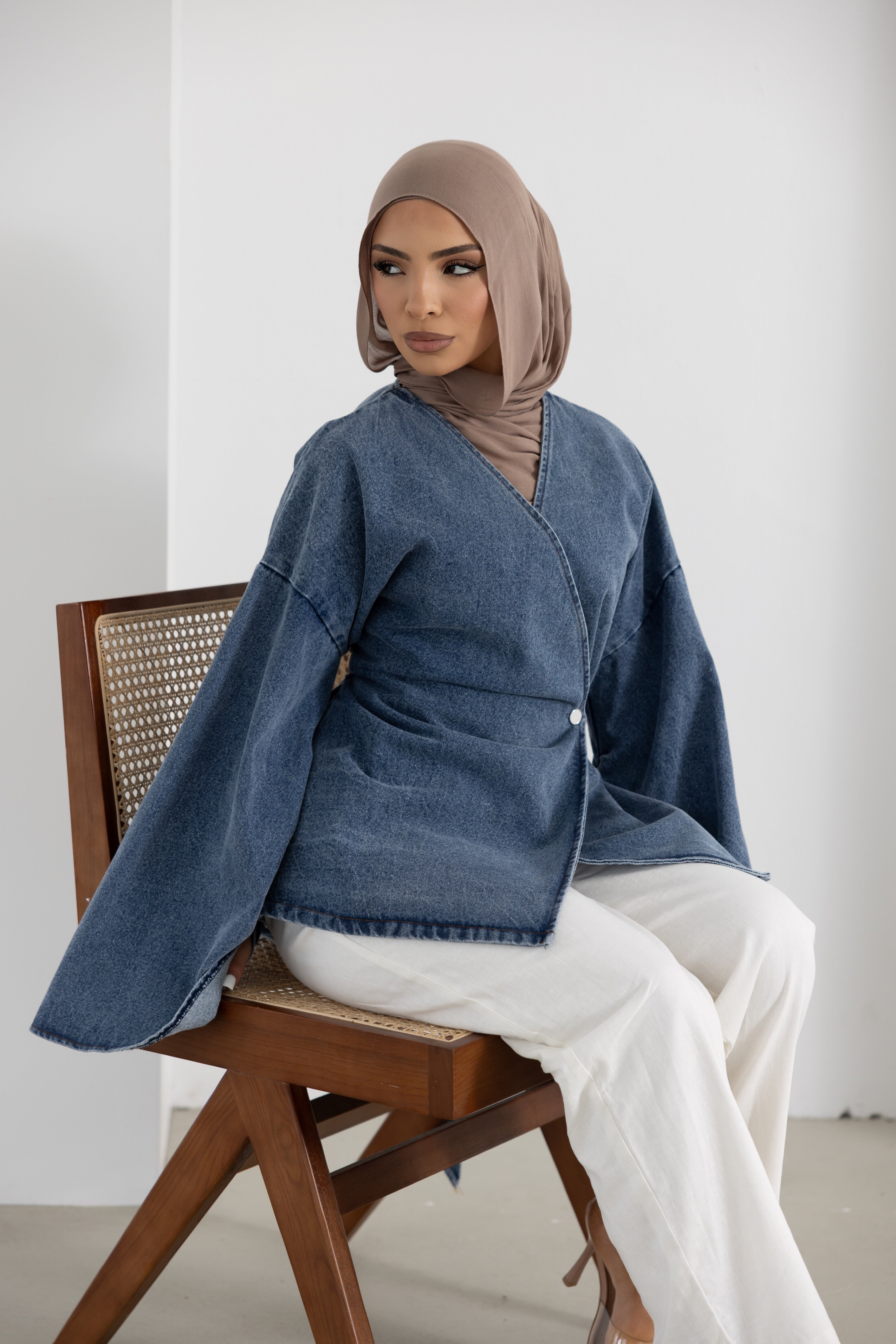 Model seated wearing the Orlena Signature Denim Mini jacket in blue denim-look 100% cotton fabric. Features a wrap-style front, wide sleeves, flattering waist-cinching fit, mini-length design, and modest silhouette. Versatile women's denim wrap jacket ideal for layering with wide-leg pants, skirts, and everyday modest fashion outfits.
