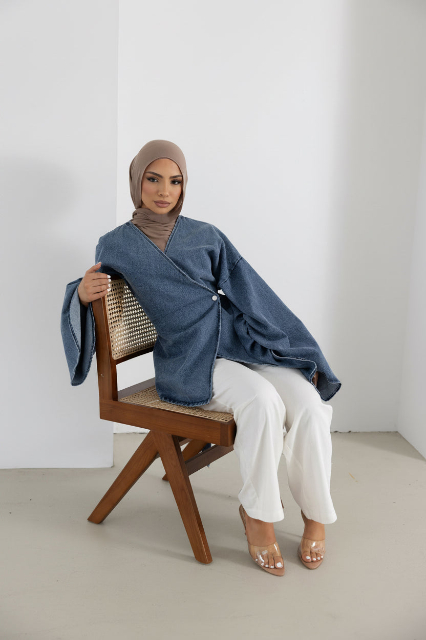 Model seated in the Orlena Signature Denim Mini jacket crafted from 100% cotton with a soft denim-look finish. Features a wrap-style front, wide statement sleeves, mini-length silhouette, and adjustable back tie waist for a flattering fit. Styled with white wide-leg pants and clear heels, creating a refined modest fashion look perfect for layering and everyday wear.