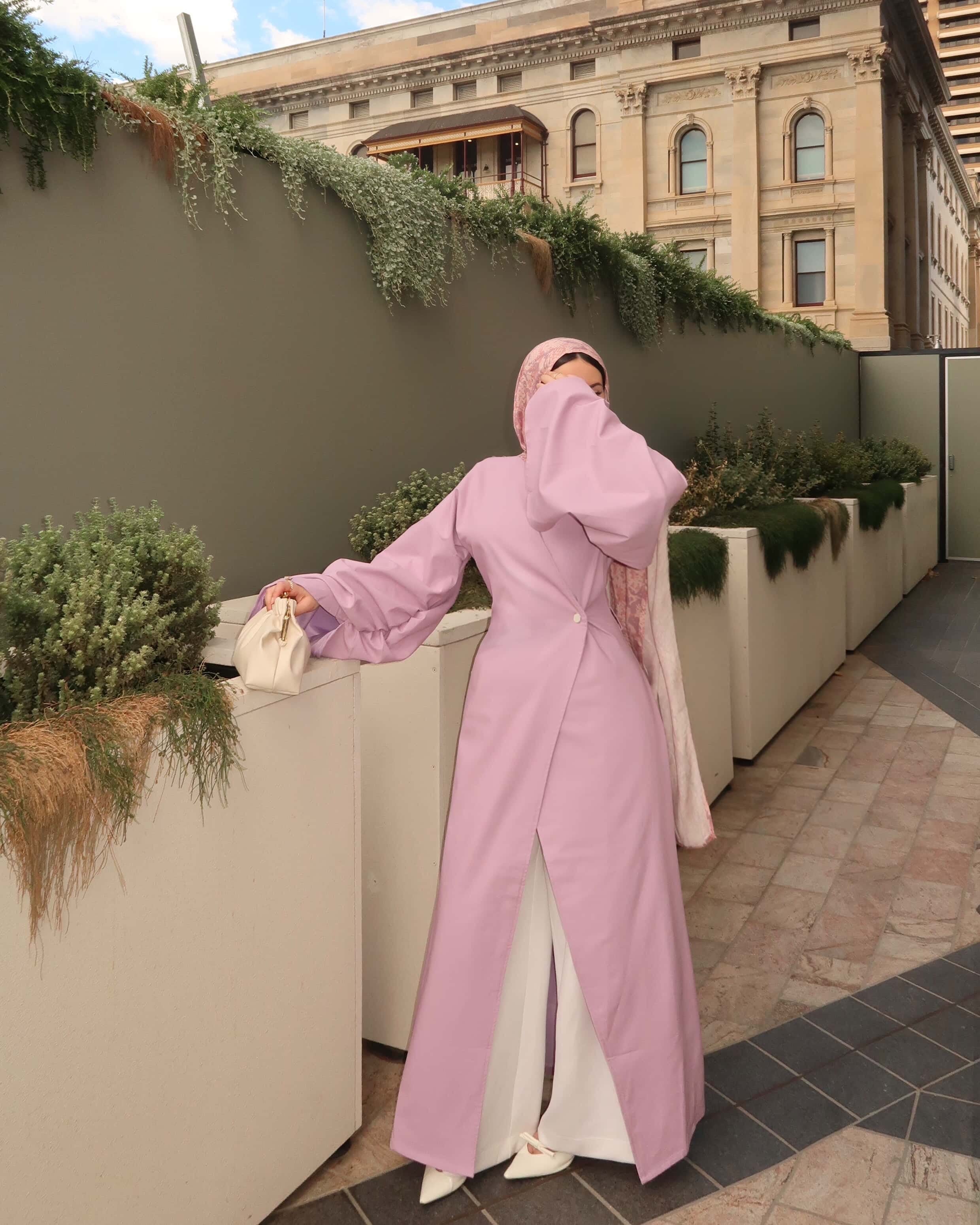 Woman wearing a pink modest wrap dress with patterned hijab and white pants styled as an elegant outdoor modest fashion outfit