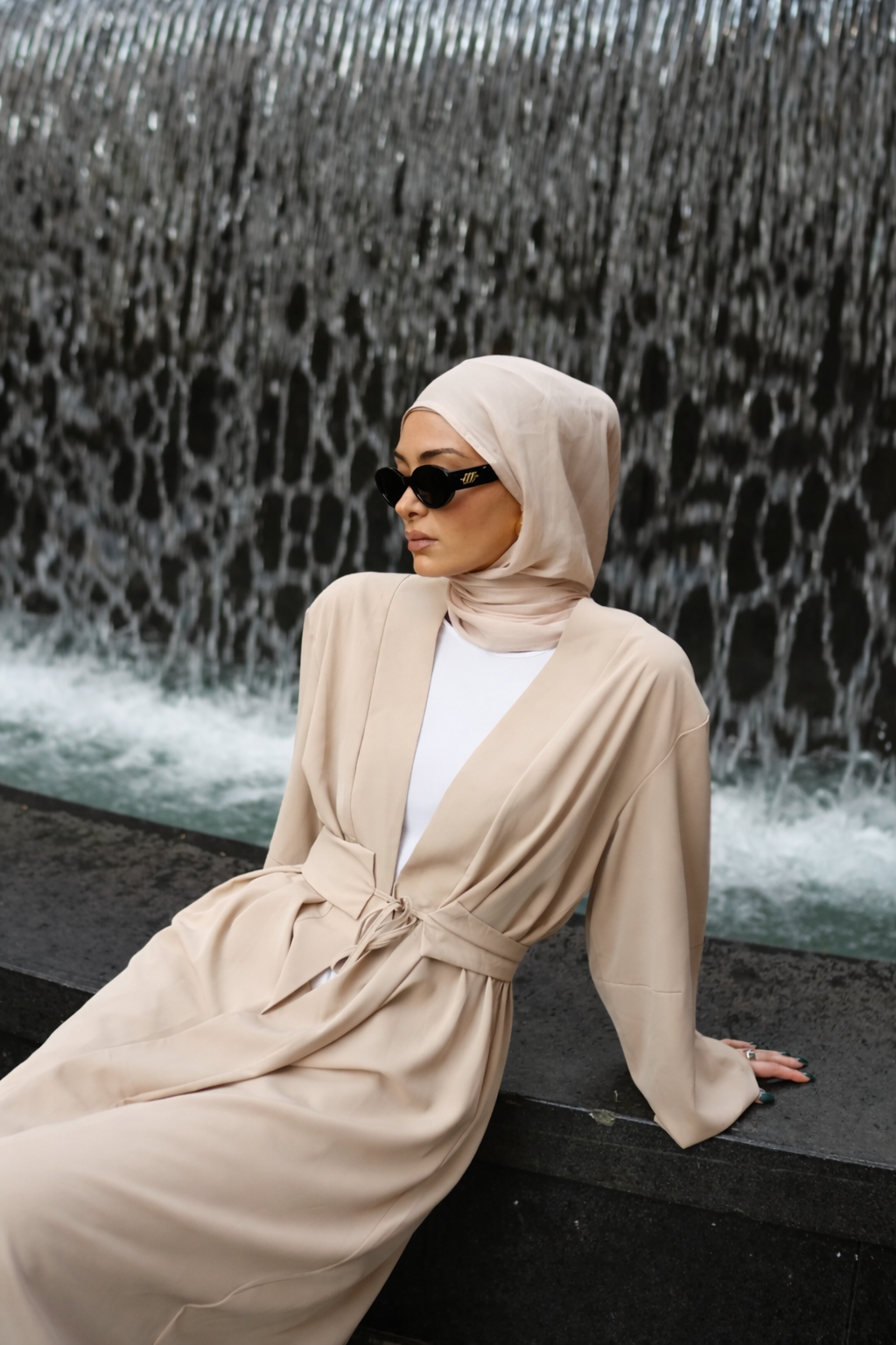 Rowan Belted Abaya in beige by Orlena Official, lifestyle shot of a modest dress with a belted waist styled with a hijab near a fountain