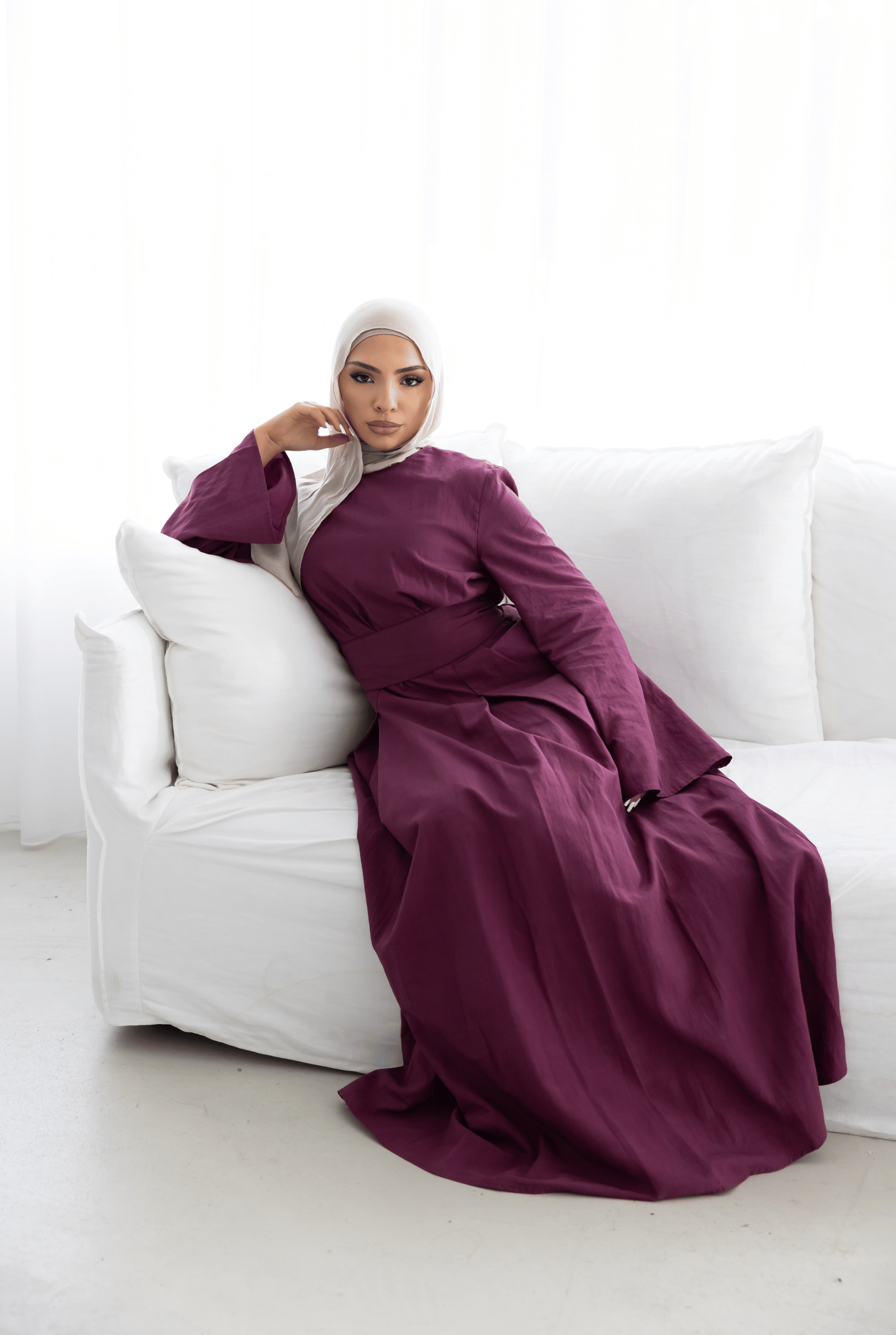 Model wearing the Sorelle Belted Dress in plum, seated in a lifestyle setting. Featuring a full-length modest silhouette, matching waist belt, wide sleeves, and breathable lyocell-linen blend fabric. An elegant and timeless modest dress designed for comfort, sophistication, and effortless everyday styling.