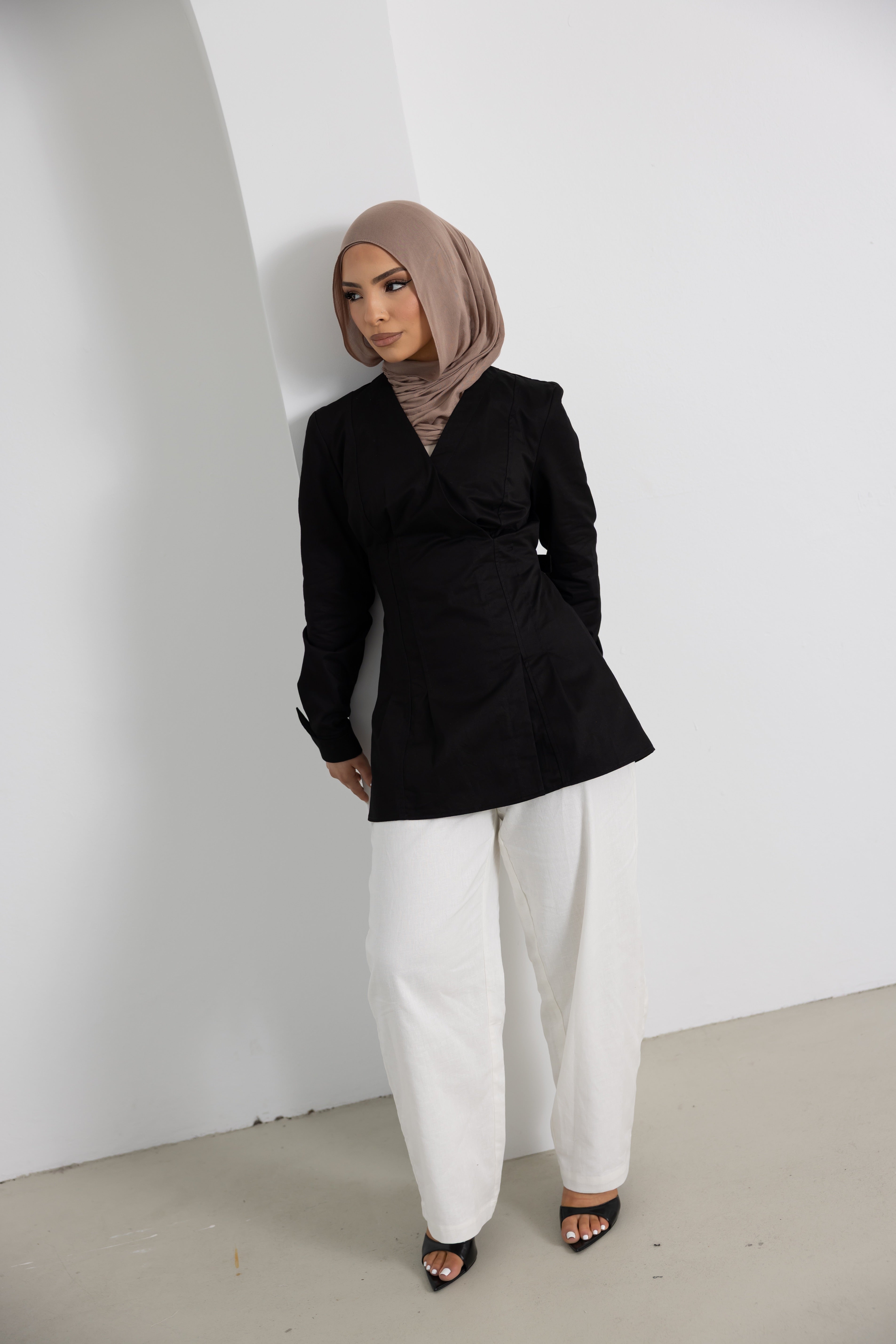 Full-length view of the Vellisse Top in black, crafted from 100% cotton with a structured yet comfortable fit. Features a flattering wrap-style front, long sleeves, and an adjustable back belt that gently cinches the waist for a feminine silhouette. Styled with white wide-leg pants and a neutral-toned hijab, showcasing an elegant modest fashion look suitable for workwear, casual styling, special occasions, and everyday wear.