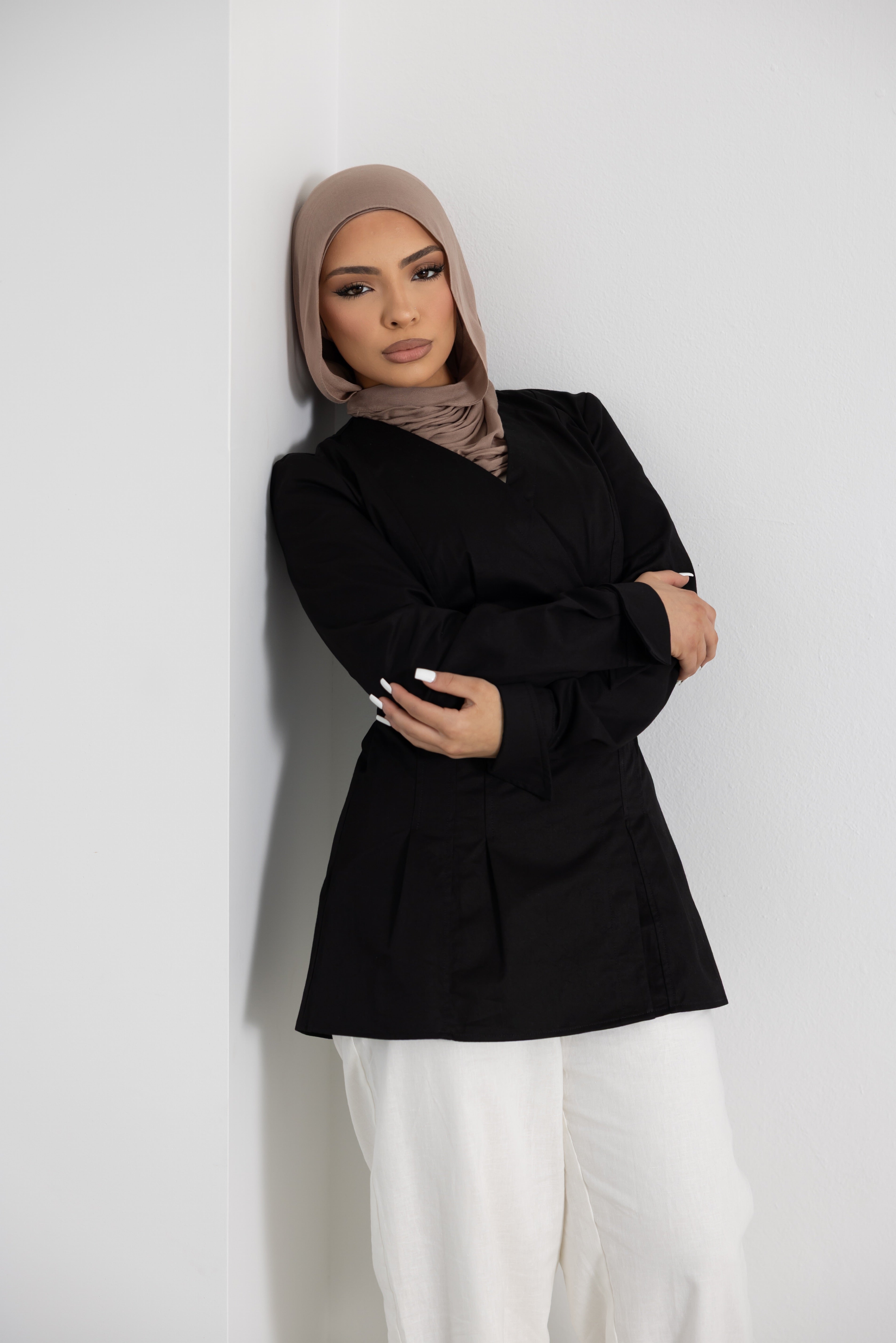 Women's Vellisse Top in black crafted from 100% cotton, featuring a flattering wrap-style front, long sleeves, and an adjustable back belt for a softly defined waist. Styled with white wide-leg pants and a neutral-toned hijab, showcasing the structured yet comfortable fit. Elegant modest fashion piece suitable for workwear, casual styling, special occasions, and elevated everyday outfits.