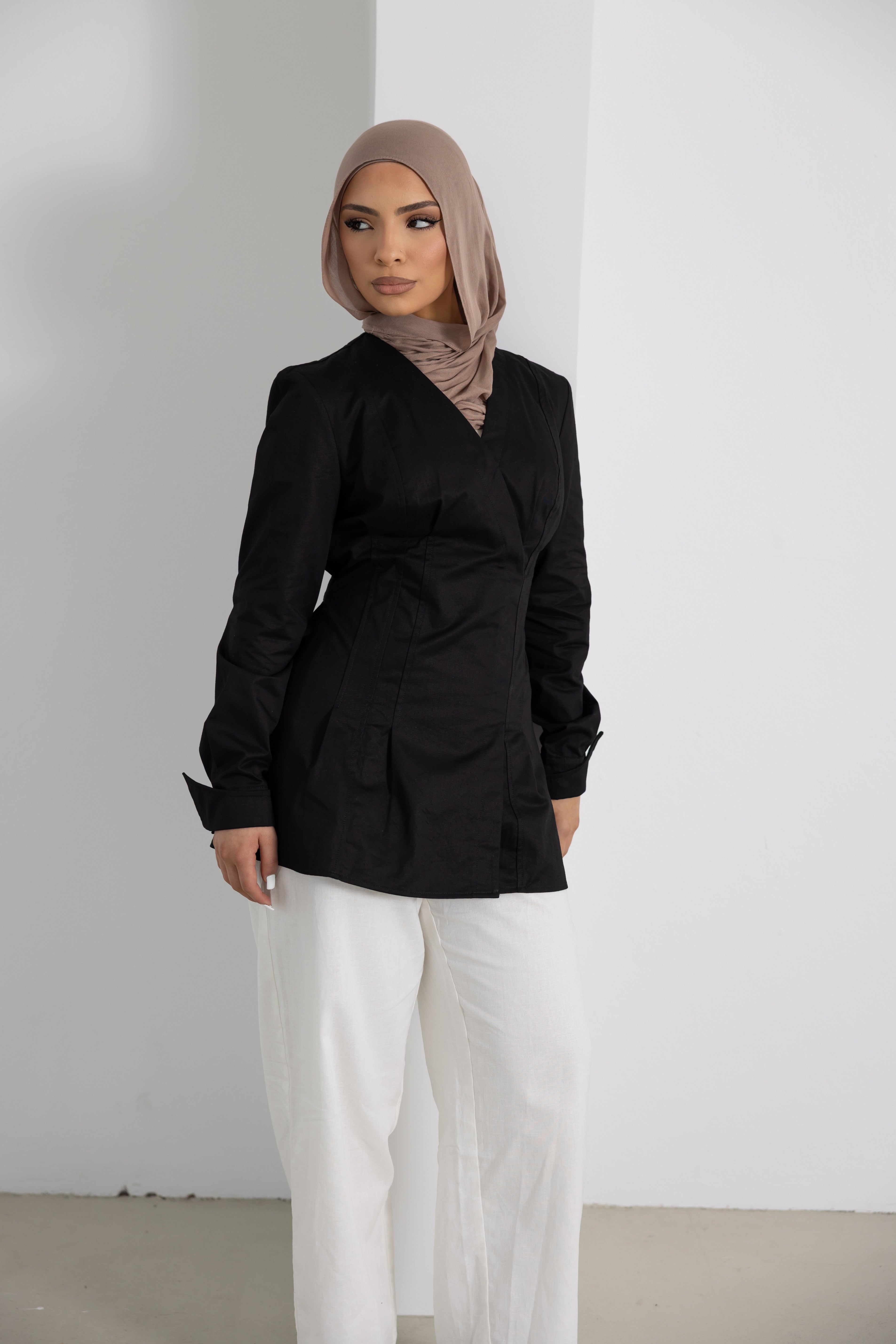 Women's Vellisse Top in black featuring a flattering wrap-style front, long sleeves, and an adjustable back belt that gently defines the waist. Made from 100% cotton with a structured yet comfortable fit, creating an elegant tailored silhouette. Styled with white wide-leg pants and a neutral hijab, showcasing a versatile modest fashion piece ideal for workwear, casual outings, special occasions, and elevated everyday dressing.