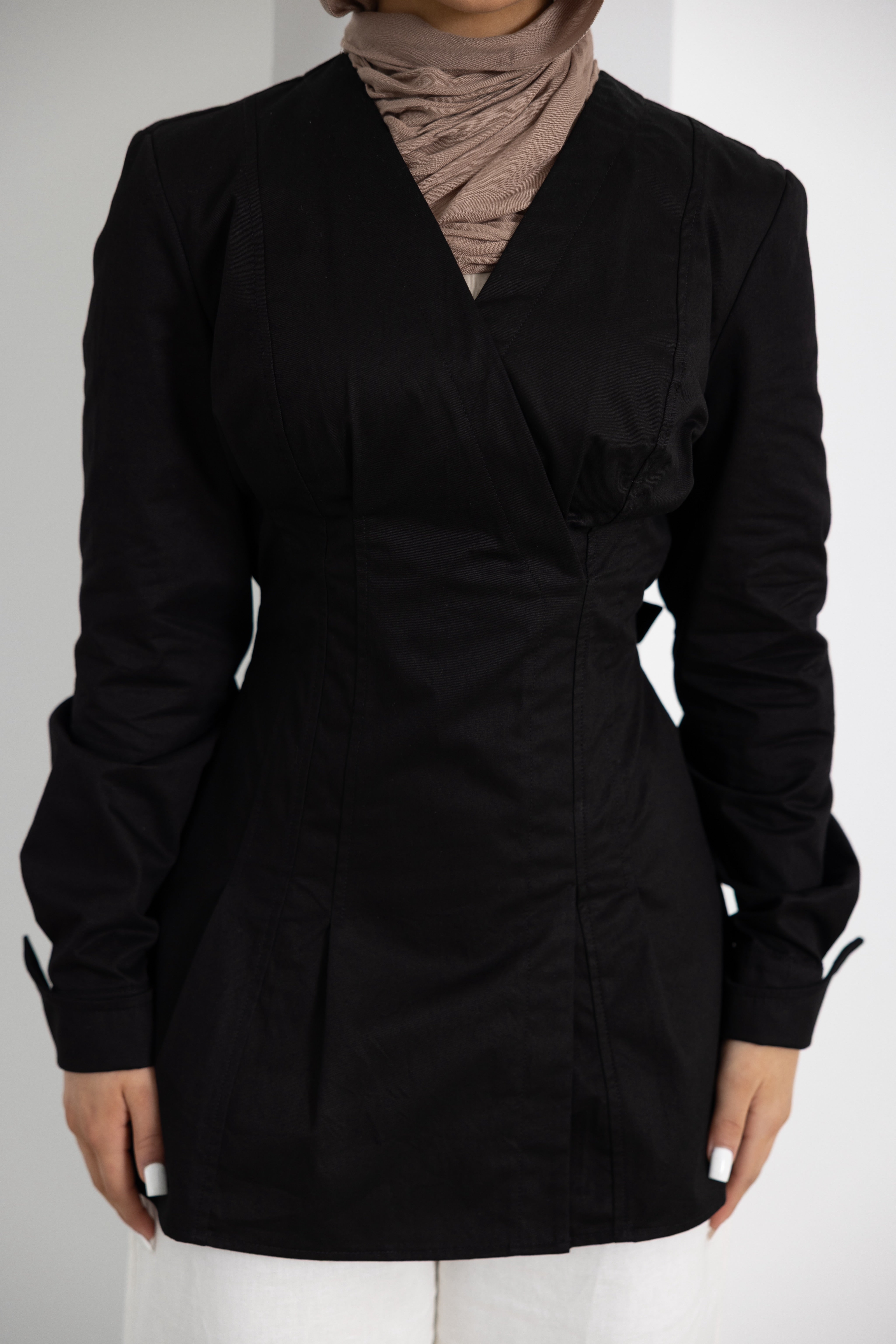 Close-up front view of the Vellisse Top in black, featuring a flattering wrap-style front, structured tailoring, and long sleeves with adjustable cuff detailing. Crafted from 100% cotton for a comfortable yet polished fit, designed to gently define the waist and create an elegant silhouette. Styled with white wide-leg pants and a neutral-toned hijab, showcasing the versatile modest design suitable for both everyday wear and elevated occasions.