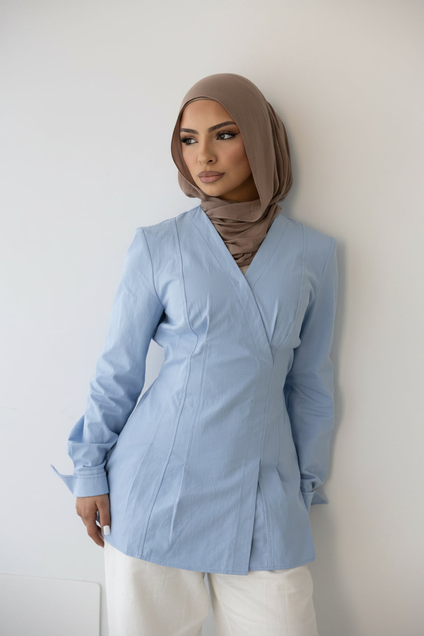 Close-up front view of the Vellisse Top in blue, crafted from 100% cotton with a structured yet comfortable fit. Features a flattering wrap-style front, long sleeves, and an adjustable back belt that gently cinches the waist for a feminine silhouette. Styled with white wide-leg pants for an elegant modest fashion look, perfect for casual wear, workwear, and elevated everyday styling.