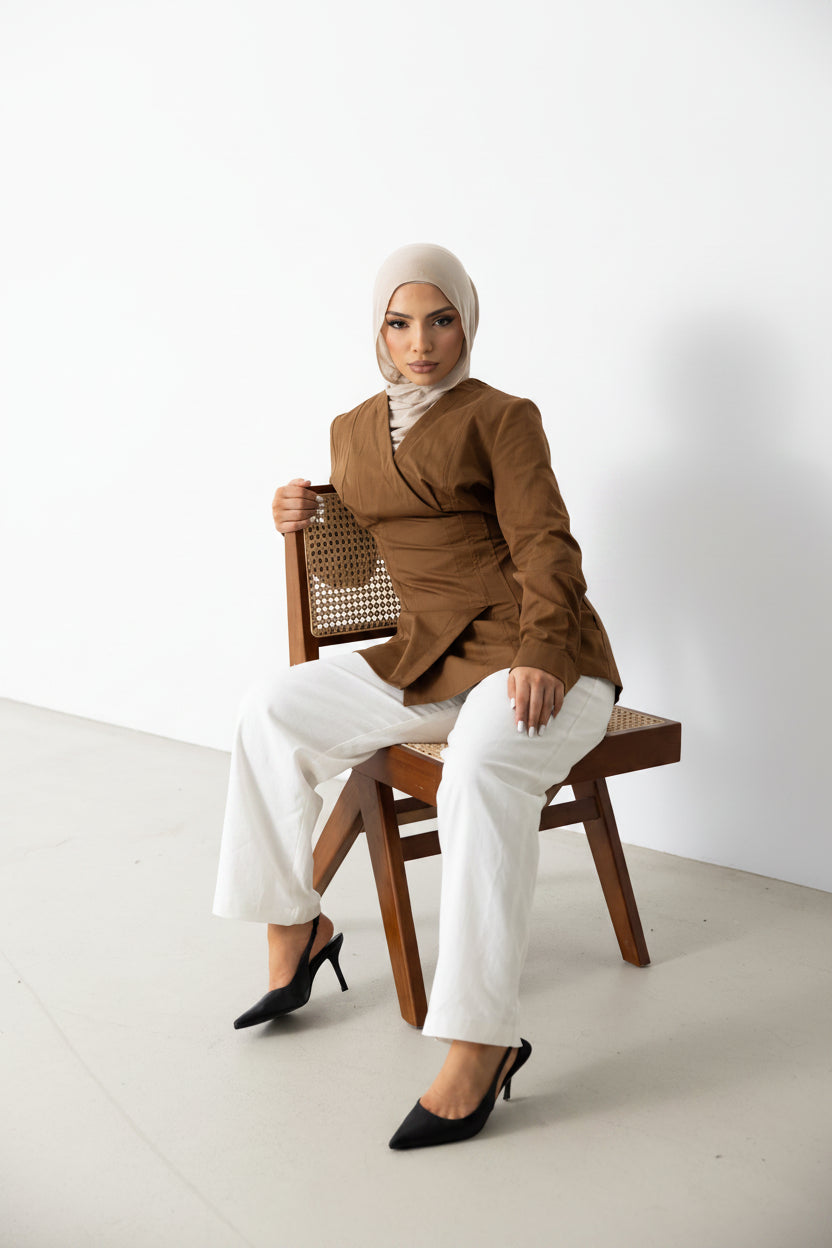 Model seated wearing the Vellisse Top in brown, crafted from 100% cotton with a structured yet comfortable fit. Features a flattering wrap-style front, long sleeves, and an adjustable back belt that gently defines the waist. Styled with white wide-leg pants and black heels, creating an elegant modest fashion look suitable for workwear, casual outings, and elevated everyday styling.